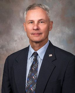 A formal headshot of Dr. Francis O’Connor, wearing a dark suit and a tie featuring the Uniformed Services University logo, against a mottled gray background.
